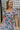 close upper front view of model wearing the Wild Blooms Midi Dress that has black fabric with a purple, pink, and blue floral pattern, ruffled hem, a square neck, a smocked back, and short puff sleeves.