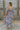 Full body back view of model wearing the Wild Blooms Midi Dress that has black fabric with a purple, pink, and blue floral pattern, ruffled hem, a square neck, a smocked back, and short puff sleeves.