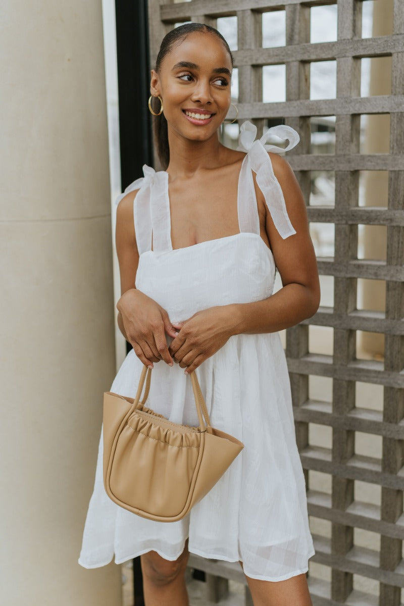 Front view of model wearing the Simple Melody Dress which features white sheer fabric with a monochromatic checkered pattern, white lining, babydoll style, a smocked back, a square neckline, organza straps with ties, and a back hook and zipper closure.