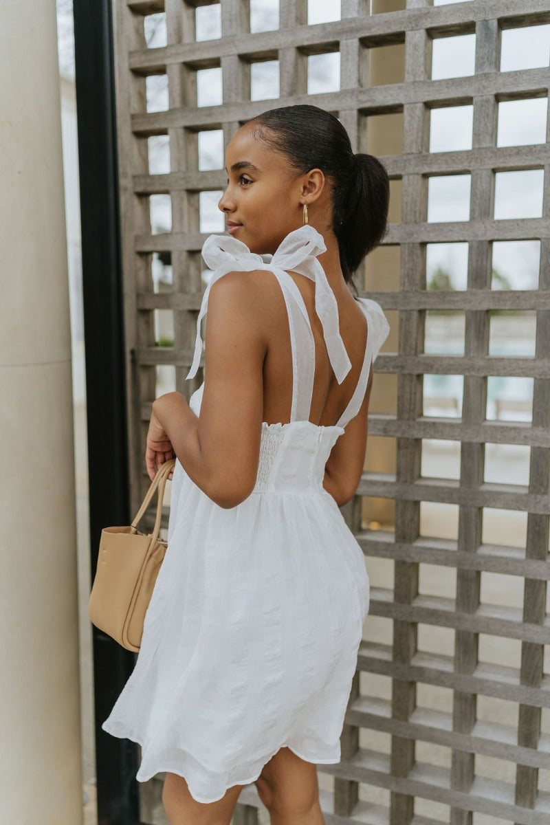 Side view of model wearing the Simple Melody Dress which features white sheer fabric with a monochromatic checkered pattern, white lining, babydoll style, a smocked back, a square neckline, organza straps with ties, and a back hook and zipper closure.