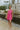 Full body view of model wearing the You Have My Heart Dress which features hot pink fabric with a lining, a diagonal ruffle and pleated skirt, a halter neckline that ties around the neck, an open back, and a monochromatic side zipper.
