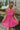 Front view of model wearing the You Have My Heart Dress which features hot pink fabric with a lining, a diagonal ruffle and pleated skirt, a halter neckline that ties around the neck, an open back, and a monochromatic side zipper.