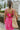 Back view of model wearing the You Have My Heart Dress which features hot pink fabric with a lining, a diagonal ruffle and pleated skirt, a halter neckline that ties around the neck, an open back, and a monochromatic side zipper.