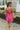 Back view of model wearing the You Have My Heart Dress which features hot pink fabric with a lining, a diagonal ruffle and pleated skirt, a halter neckline that ties around the neck, an open back, and a monochromatic side zipper.