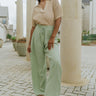 Full body view of model wearing the In Paradise Pants in Sage which features sage gauze fabric, an elastic waistband with drawstring ties, and flared legs.