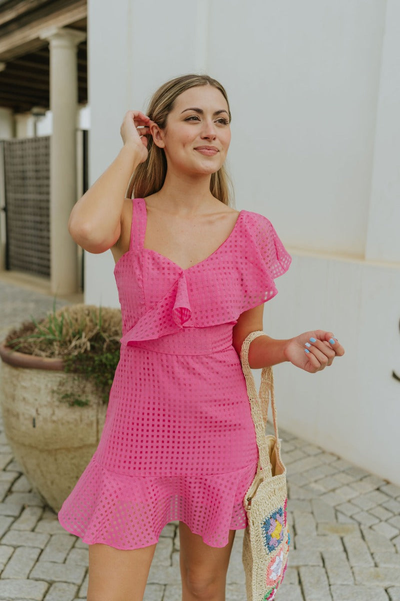Close up view of model wearing the In the Moment Dress in Pink which features pink sheer fabric with a monochromatic checkered pattern, pink lining, a ruffle hem, a sweetheart neckline with ruffle details, a thick strap and a ruffled short sleeve, and a b