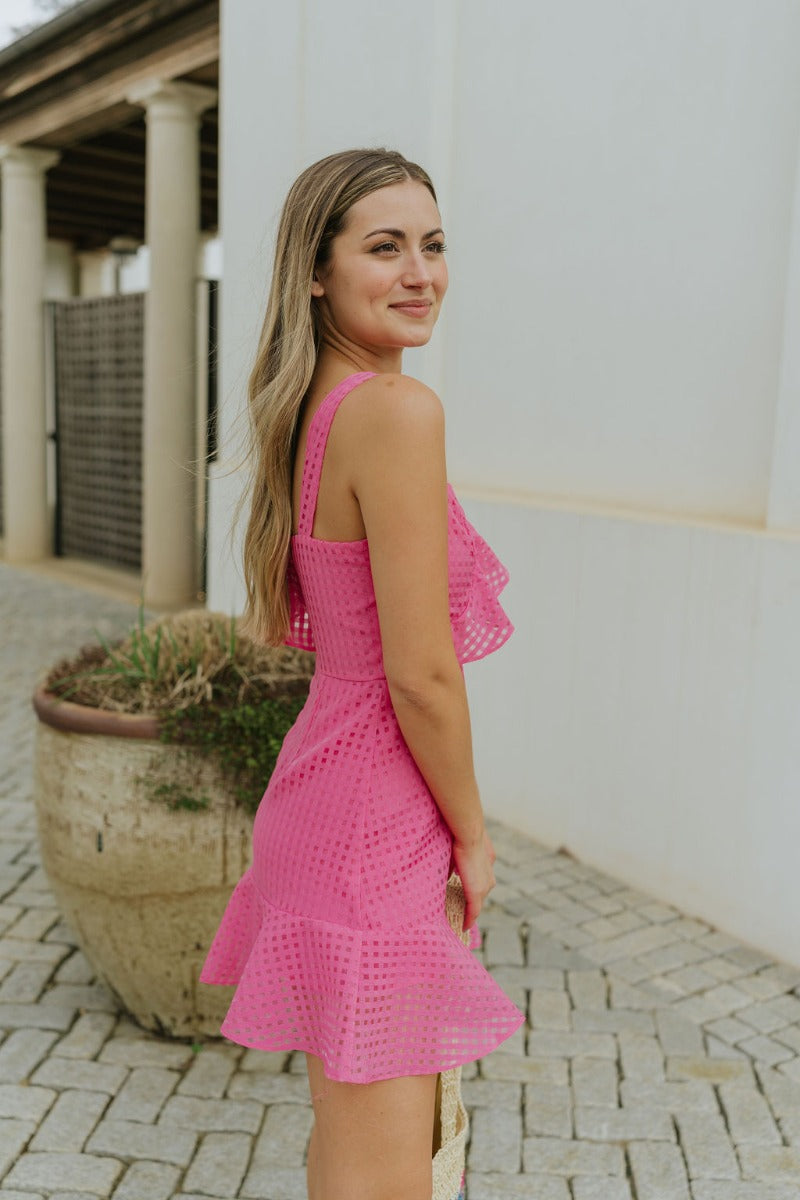 Side view of model wearing the In the Moment Dress in Pink which features pink sheer fabric with a monochromatic checkered pattern, pink lining, a ruffle hem, a sweetheart neckline with ruffle details, a thick strap and a ruffled short sleeve, and a back 
