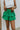 Front view of model wearing the Lucky You Skort which features green fabric with a tiered design, a mini length, green shorts lining, and an elastic waistband with ties.