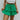 Front view of model wearing the Lucky You Skort which features green fabric with a tiered design, a mini length, green shorts lining, and an elastic waistband with ties.