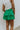 Side view of model wearing the Lucky You Skort which features green fabric with a tiered design, a mini length, green shorts lining, and an elastic waistband with ties.