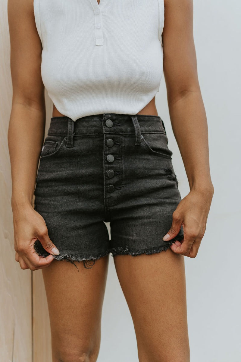Front view of model wearing the Just USA Denim: Dream On Shorts in Washed Black which features washed black denim fabric, a high-rise waist with belt loops, a five-button fly with black buttons, two front pockets, two back pockets, distressed detailing, a