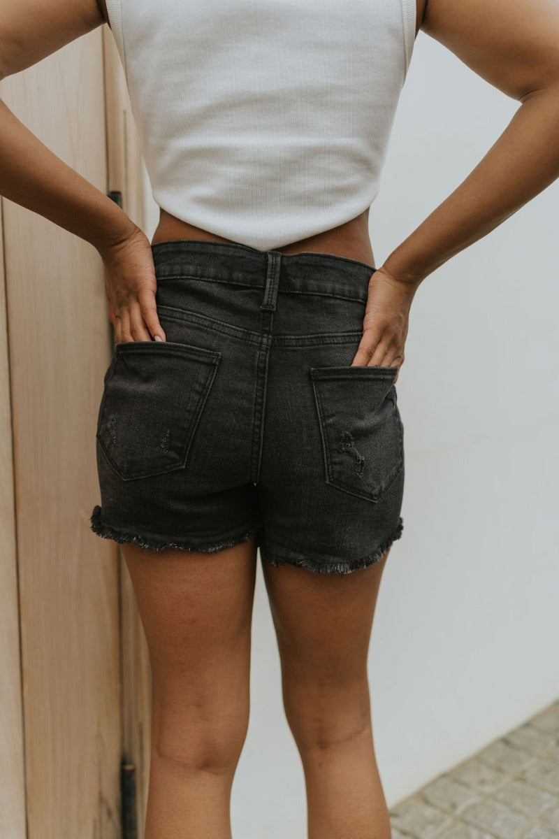 Back view of model wearing the Just USA Denim: Dream On Shorts in Washed Black which features washed black denim fabric, a high-rise waist with belt loops, a five-button fly with black buttons, two front pockets, two back pockets, distressed detailing, an