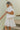 Side view of model wearing the Love Always Dress that has white fabric with a monochromatic floral pattern, white lining, a ruffle hem, an elastic waistband, a surplice neckline, and short puff sleeves.