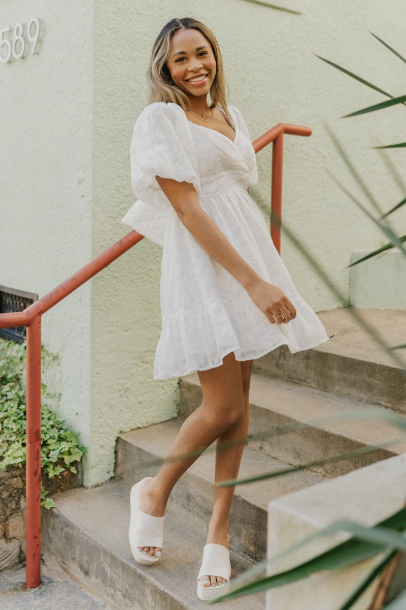Full body view of model wearing the Love Always Dress that has white fabric with a monochromatic floral pattern, white lining, a ruffle hem, an elastic waistband, a surplice neckline, and short puff sleeves.