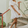 Full body view of model wearing the Love Always Dress that has white fabric with a monochromatic floral pattern, white lining, a ruffle hem, an elastic waistband, a surplice neckline, and short puff sleeves.