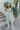 Full body view of model wearing the On The Lookout Pants which features light sage fabric, a hidden front zipper with a tortoise button closure, an elastic back waistband, two front pockets and flared legs.