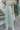 Front view of model wearing the On The Lookout Pants which features light sage fabric, a hidden front zipper with a tortoise button closure, an elastic back waistband, two front pockets and flared legs.