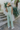 Full body/back view of model wearing the On The Lookout Pants which features light sage fabric, a hidden front zipper with a tortoise button closure, an elastic back waistband, two front pockets and flared legs.