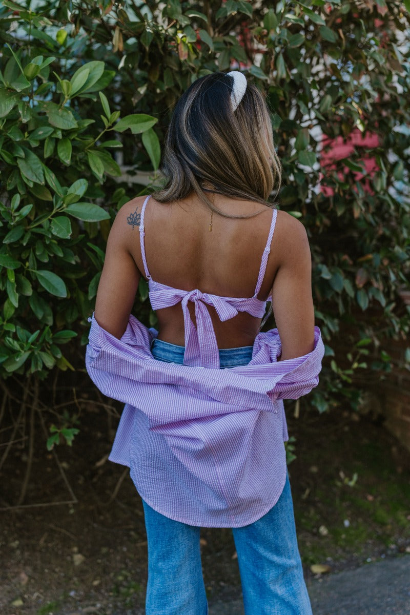 Back view of model wearing the Laguna Seersucker Bralette in Pink that has a pink and white striped seersucker pattern, adjustable spaghetti straps, and a back tie closure