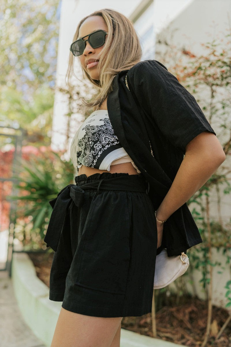 Side view of model wearing the Modern Romance Shorts which features black fabric, a high-rise elastic waistband, a tie around the waistline, two front pockets, and black shorts lining.