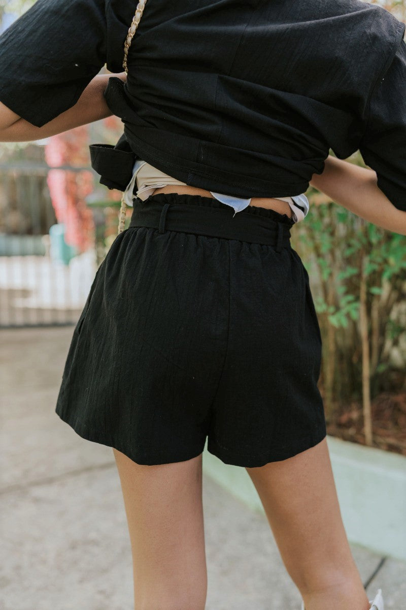 Back view of model wearing the Modern Romance Shorts which features black fabric, a high-rise elastic waistband, a tie around the waistline, two front pockets, and black shorts lining.