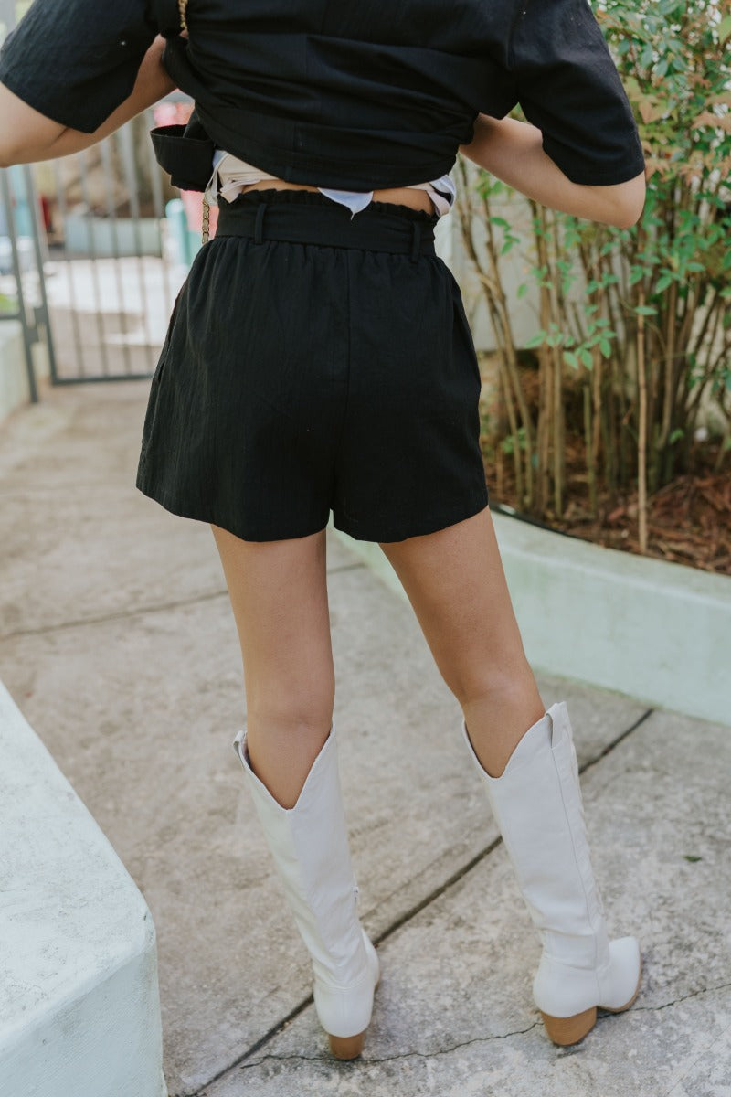 Back view of model wearing the Modern Romance Shorts which features black fabric, a high-rise elastic waistband, a tie around the waistline, two front pockets, and black shorts lining.