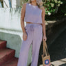 Full body front view of model wearing the Next Flight To Paradise Pants that have lavender fabric, an elastic waistband with drawstring ties, and flared legs