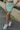 Front view of model wearing the Take Me Higher Skirt which features green and light blue knit fabric with a checkered pattern, mini length, a small front slit, and an elastic waistband.