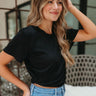 Side view of model wearing the With A Twist Top in Black that has black fabric, a cropped waist, a diagonal hem with a thick band and side twist, a round neckline, and short sleeve