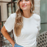 Front view of model wearing the With A Twist Top in Off White that has off white fabric, a cropped waist, a diagonal hem with a thick band and a side twist, a round neckline, and  short sleeves.