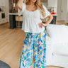 Full body front view of model wearing the Seaside Breeze Pants that have blue, light pink, yellow, green and white fabric with a palm leaf print, an elastic waistband, and wide legs
