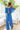 Full body back view of model wearing the Island Life Pants that have royal blue fabric with a white geometric pattern, a monochromatic side zipper with a hook closure, and wide pant legs