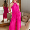Full body front view of model wearing the Valencia Jumpsuit in Magenta that has magenta fabric, an elastic waist with a tie, pockets a halter neck with a key hole, a back zipper and button closure, and flare pant legs