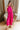 Full body front view of model wearing the Valencia Jumpsuit in Magenta that has magenta fabric, an elastic waist with a tie, pockets a halter neck with a key hole, a back zipper and button closure, and flare pant legs