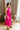 Full body side view of model wearing the Valencia Jumpsuit in Magenta that has magenta fabric, an elastic waist with a tie, pockets a halter neck with a key hole, a back zipper and button closure, and flare pant legs