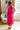 Full body back view of model wearing the Valencia Jumpsuit in Magenta that has magenta fabric, an elastic waist with a tie, pockets a halter neck with a key hole, a back zipper and button closure, and flare pant legs