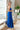 Full body side view of model wearing the Across the Ocean Maxi Dress that has royal blue fabric, maxi length, a ruffle tiered body, a tie around the waist, a smocked upper, one shoulder strap, and royal blue lining.