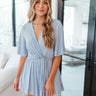 Front view of model wearing the Beach Walks Romper which features light blue fabric, elastic waistband, tie around the waist, light blue shorts lining, surplice neckline, open back with button closure, monochromatic back zipper and short flare sleeves.