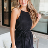 Front view of model wearing the Don't Be Shy Plisse Romper in Black which features black plisse fabric, an elastic waistband, a tie around the waist, a high neckline, thin straps, and a back button closure with a keyhole.