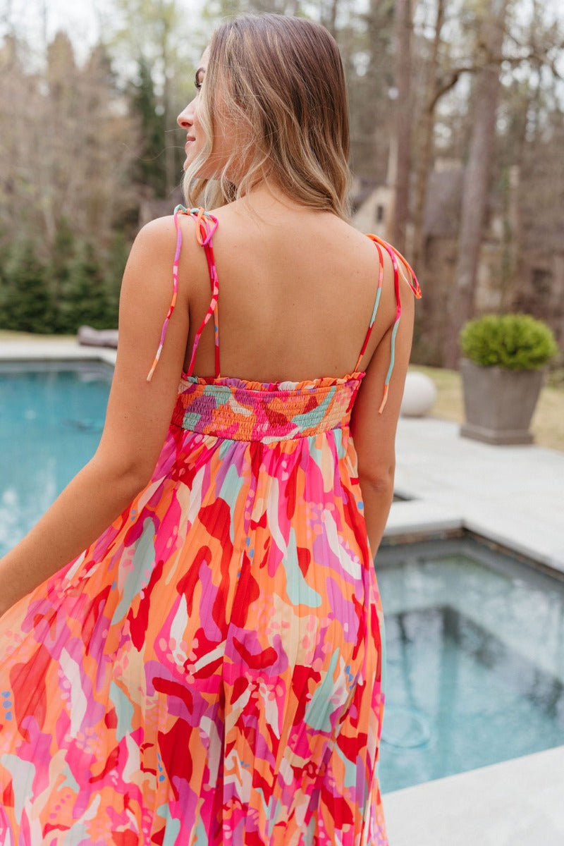 Back view of model wearing the Make Bold Moves Jumpsuit features orange, pink, red, purple and turquoise pleated fabric with a marble pattern, a smocked chest with ruffle trim, tie spaghetti straps, and peach lining.