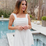Front view of model wearing the Remind Me Romper which features white fabric with white lining, a square neckline, pockets on each side, a ruched upper, thick straps, and a smocked back.