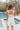 Back view of model wearing the Remind Me Romper which features white fabric with white lining, a square neckline, pockets on each side, a ruched upper, thick straps, and a smocked back.