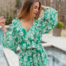 Front view of model wearing the Tropical Mindset Romper which features green fabric with a white floral print, an elastic waistband with a tie, a surplice neckline with ruffles, green lining, and long puff sleeves with elastic wrists.