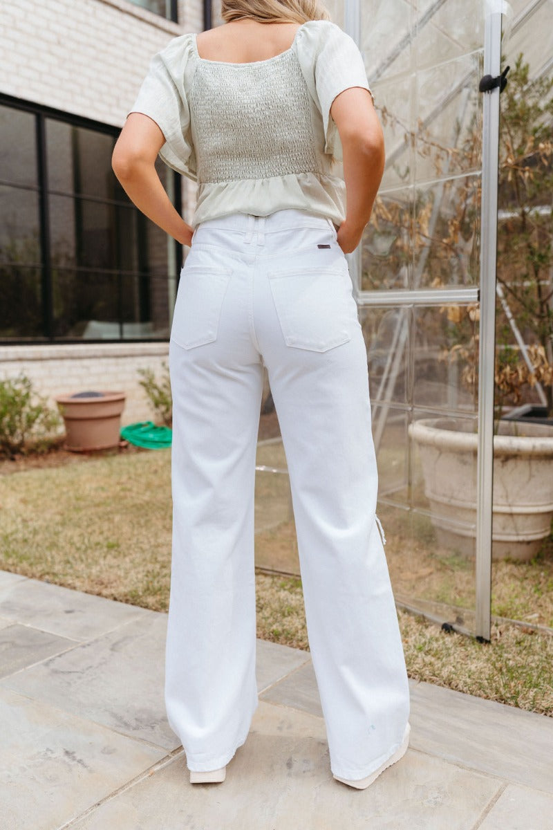 Back view of model wearing the Kancan: Where It Ends Jeans which features white denim fabric, two front pockets, two back pockets, front zipper with button closure, distressed details and wide pant leg.