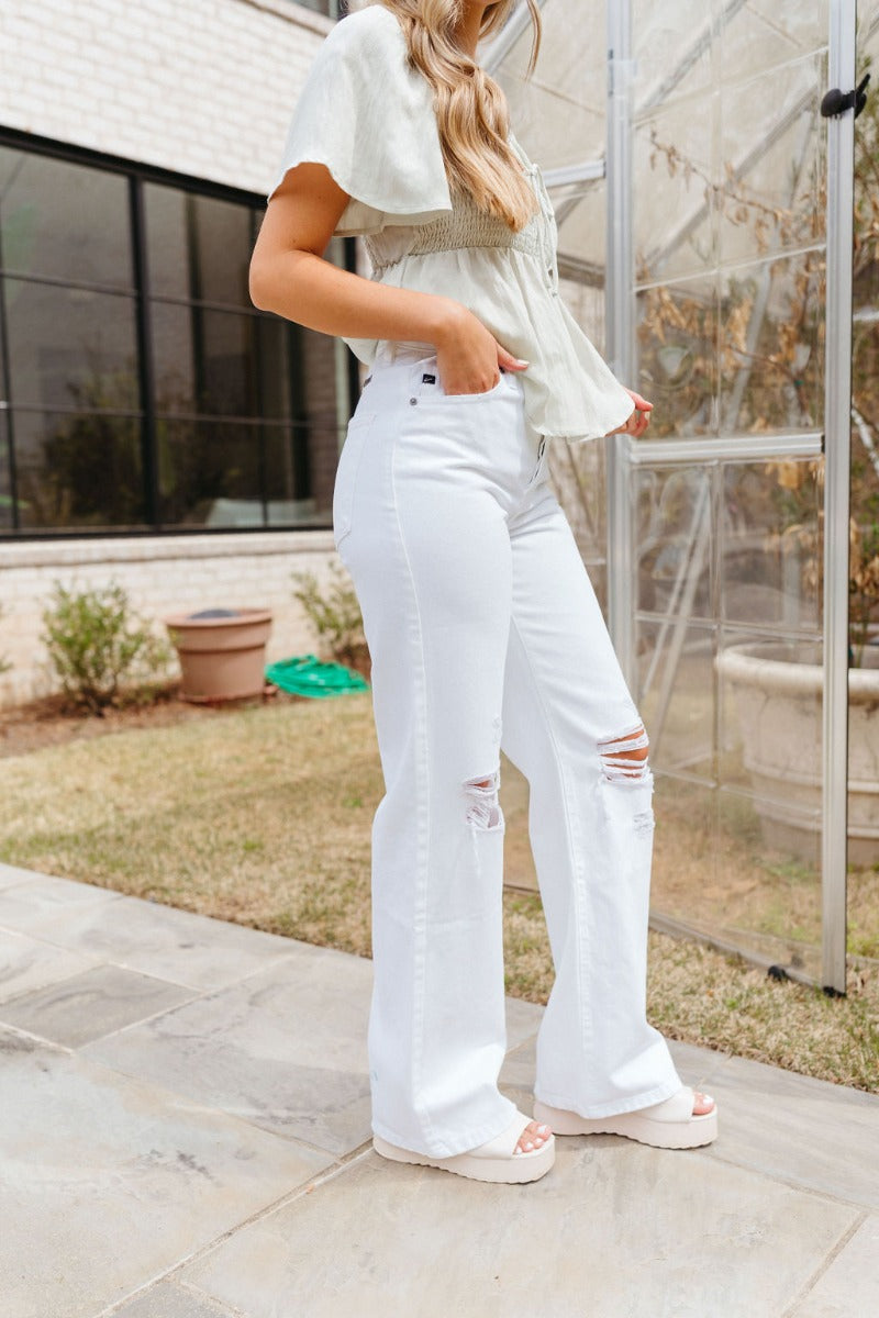 Side view of model wearing the Kancan: Where It Ends Jeans which features white denim fabric, two front pockets, two back pockets, front zipper with button closure, distressed details and wide pant leg.