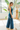 Side view of model wearing the Ready To Go Denim Jumpsuit in Indigo which features indigo denim fabric, waistband, high round neckline, sleeveless, back zipper closure and flare pant leg.