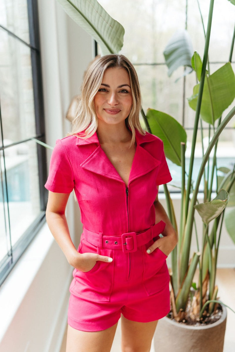 Front view of model wearing the Dakota Denim Romper in Pink that has hot pink denim fabric, front pockets, a hidden front zipper, a collared neckline, a monochromatic belt at the waistline, and short sleeves