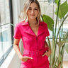 Front view of model wearing the Dakota Denim Romper in Pink that has hot pink denim fabric, front pockets, a hidden front zipper, a collared neckline, a monochromatic belt at the waistline, and short sleeves
