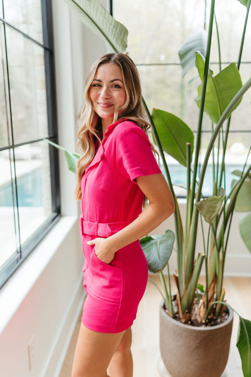 Side view of model wearing the Dakota Denim Romper in Pink that has hot pink denim fabric, front pockets, a hidden front zipper, a collared neckline, a monochromatic belt at the waistline, and short sleeves