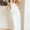 Full body view of model wearing the Go With The Flow Jumpsuit which features off white fabric, an elastic waistband, a round neckline, an upper overlay, side pockets, off white lining, a button back closure, and flared pant legs.